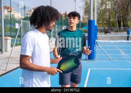 Caucasian young male instructor talking to a young african man playing pickleball in an outdoor court during sunny day Stock Photo