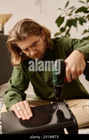 Young man in eyeglasses sitting on the floor and bending over plastic bucket while making several holes with electric handtool Stock Photo