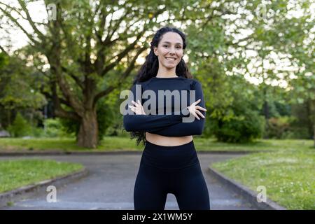 A cheerful Hispanic woman in athletic attire confidently poses with crossed arms in a lush park, showcasing a fit and healthy lifestyle. Stock Photo