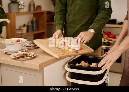 Hands of young unrecognizable man standing by kitchen counter and throwing leftover food in compost after cooking vegetable stew Stock Photo