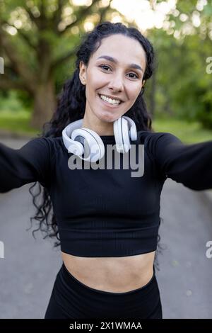 A smiling Hispanic woman enjoys a workout break in the park, taking a selfie with her headphones around her neck. She wears a black sports outfit. Stock Photo