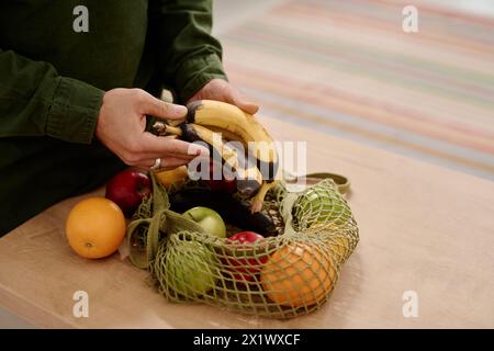 Hands of young unrecognizable man holding bunch of rotten bananas over shopping bag with fresh ripe apples and oranges on table Stock Photo