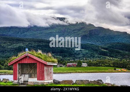 Quaint red bus shelter with a lush green sod roof stands by Halsafjord against a backdrop of forested mountains and dramatic clouds in summer, Norway Stock Photo