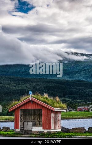 Traditional red bus shelter with a green sod roof stands before a mountainous dramatic cumulus clouds backdrop in the village Halsa, Trøndelag Norway Stock Photo