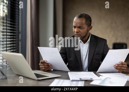 Sceptic formal male in business suit looking attentively on sheets of paper in both hands while sitting by desktop. Focused accountant comparing data on two documents from different departments. Stock Photo