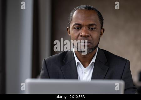Portrait of concerned diverse man looking attentively into monitor of computer on blurred background. Strict employer in shirt checking productivity of marketing department remotely from cabinet. Stock Photo