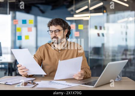 Thoughtful financial advisor in eyeglasses checking and analyzing printed graphs on paper sheets in personal workplace. Attentive man reading additional data for making deep research of investments. Stock Photo