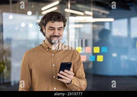 Carefree guy with stylish hairstyle using black cell phone while standing alone in working space. Relaxed man watching interesting video in social media while using wireless internet connection. Stock Photo