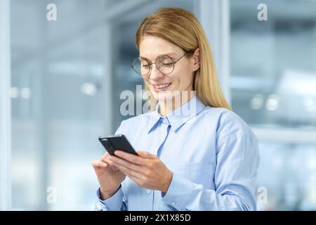 Portrait of attractive blonde woman in round eyeglasses looking down at cell phone in hands and smiling. Caucasian female making break from business and checking social media through digital gadget. Stock Photo