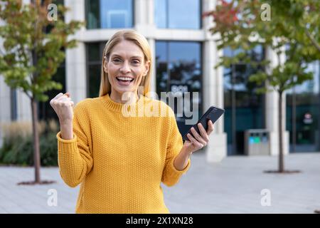 Happy mature woman in a bright yellow sweater celebrates success while holding a smartphone, outdoor setting with modern architecture in the background. Stock Photo