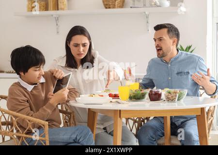 Teenage boy using tablet computer and his angry parents at table during dinner in kitchen. Family problem concept Stock Photo