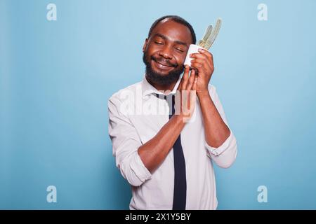 Young plant lover with eyes closed white pot with cactus to cheek showing love for houseplant. Smiling african american office worker with caring personality holding a potted plant. Stock Photo