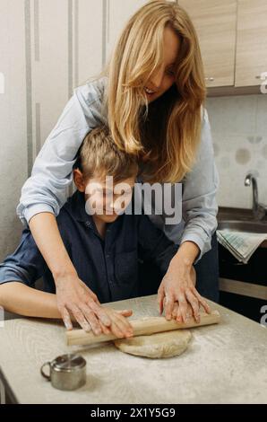 A woman and a boy are seen in a kitchen setting, engaging in the process of making pizza dough. The woman is guiding the boy as they knead the dough, Stock Photo