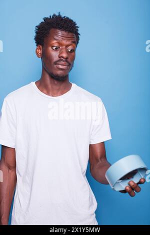 Close-up of african american man carrying wall clock with shocked expression. Portrait of black male individual with surprised face, looking at hours and minutes on a blue watch for alarm. Stock Photo