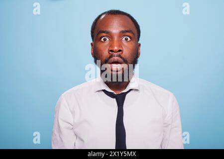 Stunned businessman stands in front of an unbelievable situation on a blue background, his mouth hanging open in shock. Surprised black individual with wide eyes and a jaw drop. Stock Photo