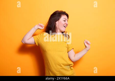 Cheerful woman happily dancing with hands in the air in front of isolated background. Caucasian lady radiating positivity, showcasing dance skills, and enjoying herself during casual photo shoot. Stock Photo