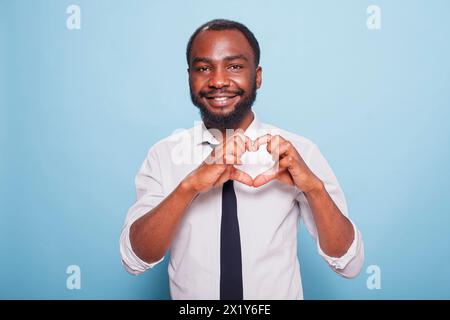 Smiling african american influencer making heart shape fingers to show care for supporting fans. Joyful male blogger in white shirt and black tie doing the hand gesture for love on his chest. Stock Photo