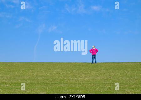 Cyclist with Helmet and Pink Windbreaker Standing Alone on Dike in Ostfriesland Under Bright Blue Sky. green, pink, bicycle helmet, outlook, dike, sun Stock Photo