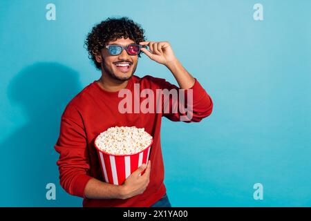 Photo of latin guy in red sweatshirt touch 3d goggles and laughing watching comedy movie with popcorn isolated on cyan color background Stock Photo