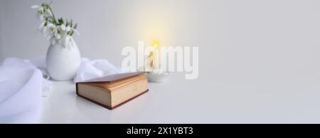 panorama, banner of still life in high key style, old paper book in a dark red binding on white table, candle, bouquet of half snowdrops in vase, conc Stock Photo