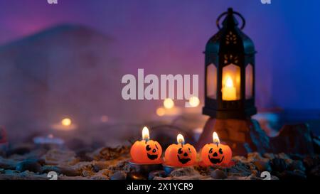 Halloween background, 3 pumpkin-shaped candles in the foreground, an old lamp on a rock against a blue out-of-focus background with yellow lights and Stock Photo