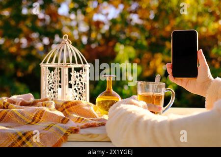 on the table in the garden tea in a glass mug, a candle is burning, a woman is holding a smartphone with a blank black screen, the concept of an outdo Stock Photo