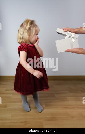 little blonde girl 2 years old in a red velvet dress stands in profile in the room, man's hands show a gift, the concept of innocence, childhood Stock Photo