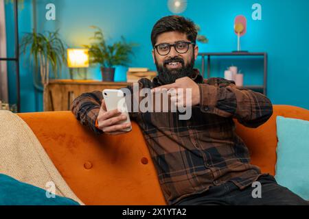 Smiling happy Indian man using smartphone application online pointing showing like thumbs up remote distance job working from home. Bearded guy freelancer recommends to use a good mobile program app Stock Photo