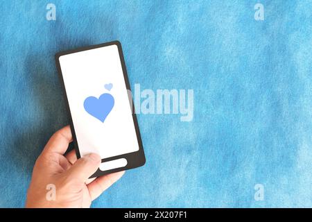 Human hand holding mobile phone with blue heart icon. Kindness, love and positivity in social media internet concept. Stock Photo