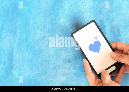 Human hand holding mobile phone with blue heart icon. Kindness, love and positivity in social media internet concept. Stock Photo