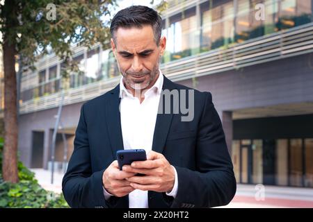 Serious businessman using an application on his smartphone and standing outdoors Stock Photo