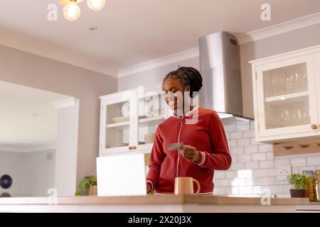 Black young woman with credit cards, standing in kitchen by an open laptop Stock Photo