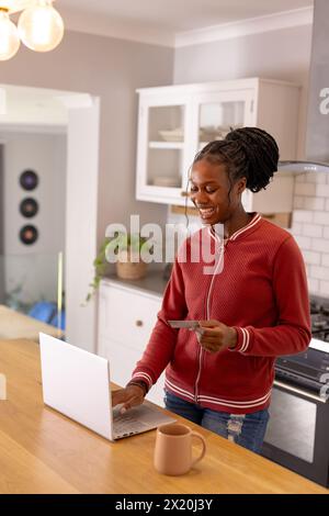 African American young woman holding credit cards, using laptop indoors in her kitchen at home Stock Photo