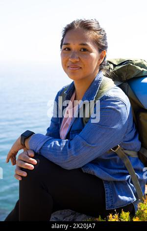 Biracial female hiker sitting, wearing a backpack, adventure in nature Stock Photo