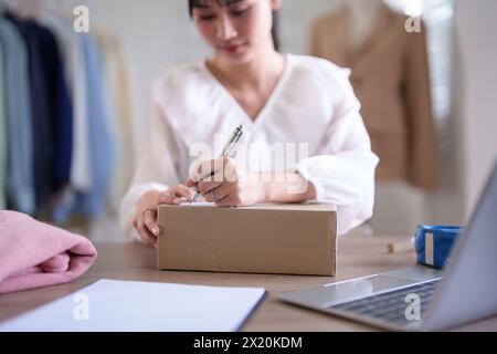 Young asian merchant women writing address data in cardboard box and preparing to shipping delivery customer while checking purchase online order on l Stock Photo