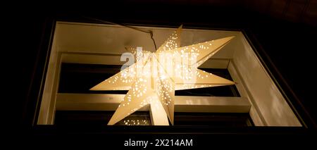 A star shaped paper lantern hangs from a window. The lantern is lit up and shining brightly. Scene is warm and inviting Stock Photo