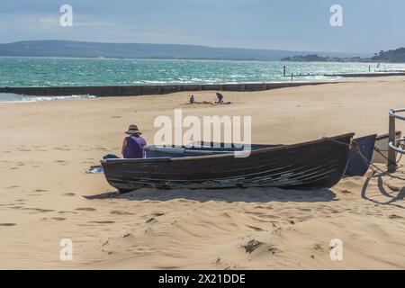 Middle Chine, Bournemouth, UK - July 12th 2023: Woman sitting on the beach behind three rowboats. Stock Photo