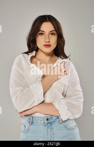 A stunning plus-size woman strikes a pose in a white shirt against a gray backdrop, exuding confidence and style. Stock Photo