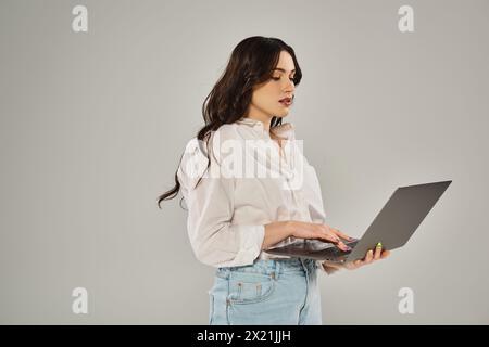 A beautiful plus size woman in stylish attire holds a laptop with confidence on a gray backdrop. Stock Photo