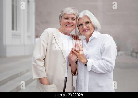 Portrait of beautiful smiling elderly senior women with gray hair embracing, dressed in stylish white clothing, mature female models together on city Stock Photo