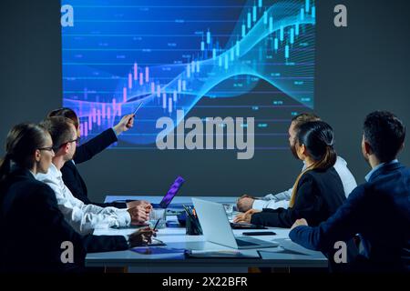 Business professionals sit around conference table in office boardroom, examining digital stock chart displayed on monitor. Stock Photo