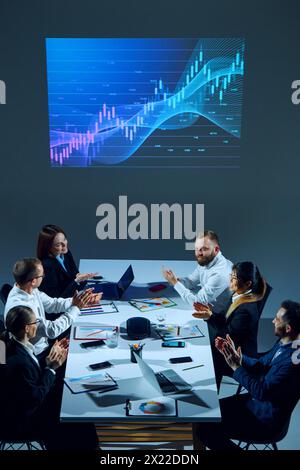Colleagues in corporate setting analyze market data while examining a digital stock chart displayed on a monitor. Stock Photo