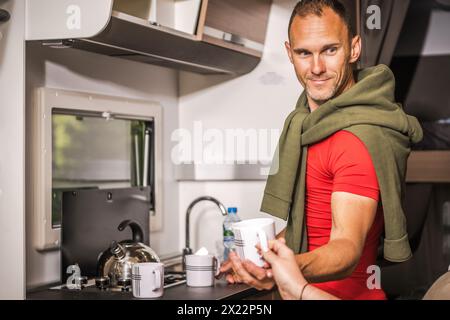 Happy Caucasian Man Serving Hot Tea While Standing Next to Camper Van Kitchen. Summer RV Road Trip Stock Photo