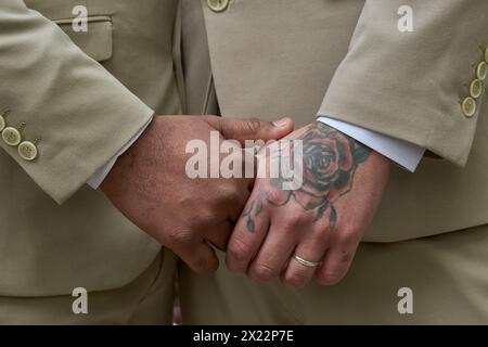 Two men in suits holding hands and one has a rose tattoo on his hand. Concept of love and unity between the two men Stock Photo