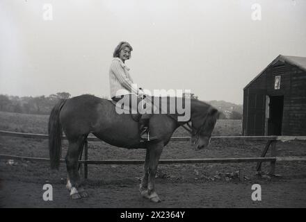 1970s, historocal, at a riding centre, a woman sitting on horse beside a fence near the stables, England, UK. Stock Photo