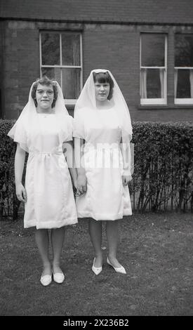 1970s, historical, two catholic schoolgirls in white dresses and wearing hair veils, Catholic standing for a photo after having their first holy communion, England, UK. Stock Photo