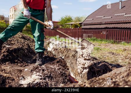 Worker covering pipes in trench with sand using shovel. Building house. Man builder wearing uniform. Construction Stock Photo
