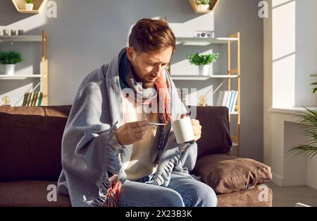 Sick young man sitting on sofa with thermometer wrapped in plaid Stock Photo