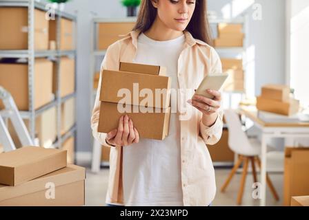 Young female employee holding cardboard boxes standing in warehouse with mobile phone. Stock Photo