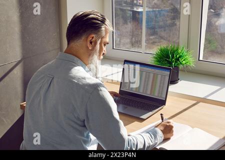 Back view of mature business man working on a laptop with tables at office and writing. Stock Photo